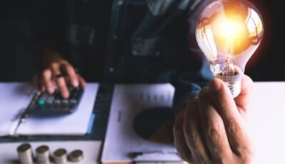 Pessoa segura uma lâmpada acesa enquanto faz cálculos financeiros, representando o impacto da bandeira vermelha na conta de energia.