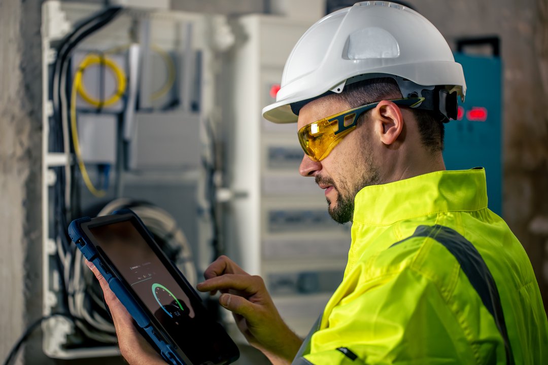 Técnico usando capacete e óculos de proteção enquanto opera um tablet para monitorar um painel elétrico, ilustrando processos de digitalização da energia.